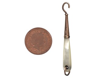 May include: An antique copper-toned button hook with a pearlescent handle, next to a copper coin. The hook has a decorative etched design near the top and a small loop at the bottom. The coin is round with a profile of a person.