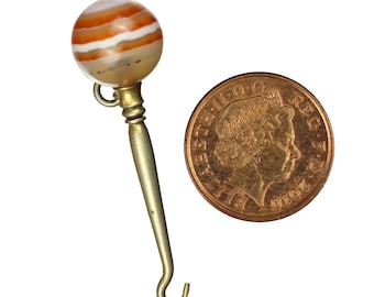 May include: A vintage rattle with a striped agate ball and a brass handle. The rattle is next to a coin for size comparison. The agate ball has bands of orange, white, and tan. The handle has a hook at the end.