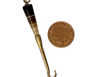 May include: An antique crochet hook with a black, brown, and white striped agate handle and a brass hook. The hook is next to a coin for size comparison. The hook's handle is approximately 1.5 inches long.