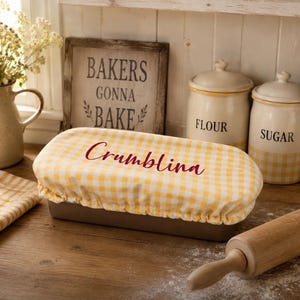 May include: A rectangular baking dish covered with a yellow and white gingham fabric. The word "Crumblina" is written across the fabric. A wooden rolling pin and two canisters labeled "Flour" and "Sugar" are also visible.