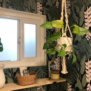 May include: A bathroom with tropical-themed wallpaper featuring large green leaves and pink flowers. A potted plant hangs from the ceiling in a macrame plant hanger. A wooden shelf with a wicker basket, a bottle of soap, and a candle is mounted on the wall.