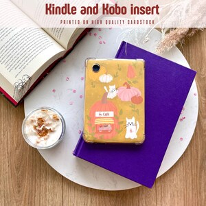 Puede incluir: Un inserto para Kindle y Kobo con un diseño de temática otoñal con calabazas, gatos y el texto "So Cafe". El inserto está encima de un libro morado y un plato blanco, con un libro abierto y una taza de café cerca.