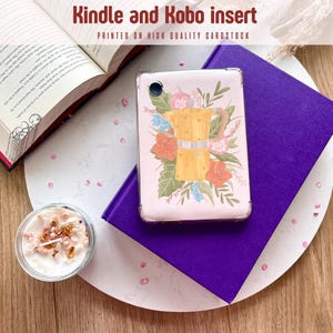 Op de afbeelding: Een Kindle- en Kobo-inzetstuk met een bloemenontwerp met een gele koffiezetter. De inzet zit op een paars boek, met een open boek en een kaars op de achtergrond. De tekst "Kindle and Kobo insert" is zichtbaar.
