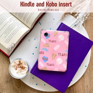 Op de afbeelding: Een roze Kindle- of Kobo-hoesje met een patroon van harten, wortels en andere afbeeldingen, met het woord "TEARS" herhaald. De hoes ligt op een paars boek en een witte schaal, met een open boek en een klein kopje eten op de achtergrond. De tekst "Kindle and Kobo insert" staat bovenaan.