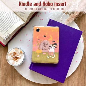 Op de afbeelding: Een Kindle- en Kobo-inzetstuk met een Halloween-thema. De inzet toont een kat met een heksenhoed, een grafsteen met de tekst "Read in Peace" en vleermuizen. De inzet ligt op een paars boek en een witte plaat.