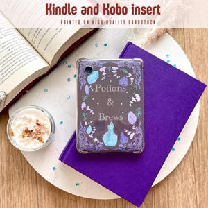 Könnte beinhalten: Ein Kindle- und Kobo-Einsatz mit dem Text "Potions & Brews" auf dunklem Hintergrund, umgeben von floralen und Kristall-Designs. Der Einsatz liegt auf einem lila Buch, zusammen mit einem offenen Buch und einer kleinen Tasse Sahne.