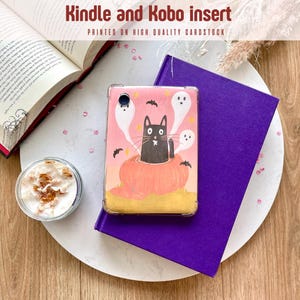 Może przedstawiać: Różowa wkładka do Kindle i Kobo z motywem Halloween. Przedstawia czarnego kota w dyni z duchami i nietoperzami. Tekst brzmi "Kindle and Kobo insert". Wkładka leży na białym talerzu z fioletową książką.