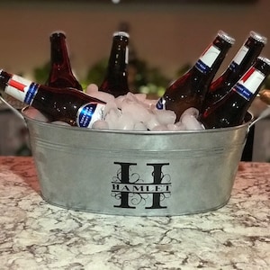 May include: A silver metal ice bucket with a black monogram "H" and the word "HAMLET" on the front. The bucket is filled with ice and six bottles of beer.