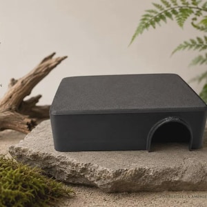 Pode incluir: Um abrigo retangular preto para répteis com uma superfície superior lisa e uma única entrada em arco. O produto apresenta etiquetas de texto incluindo SECURE HIDEAWAY, STRESS REDUCTION, EASY TO CLEAN e FOR REPTILES & AMPHIBIANS.