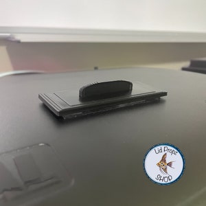 May include: Black plastic lid prop with a textured surface and a raised ridge on the top. The prop is designed to hold a lid in place.