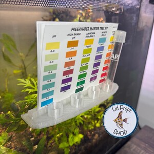 May include: A white plastic holder with test tubes for a freshwater aquarium test kit. The kit includes tests for pH, ammonia, and nitrite. The holder is attached to the side of an aquarium with a green plant in the background. The text on the kit reads "Freshwater Master Test Kit".