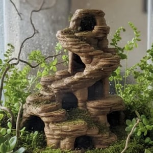 May include: A tan, multi-tiered rock-like structure with a spiral staircase design and several dark, hollow cave openings. The structure is surrounded by small green ferns and succulent plants, creating a layered, naturalistic appearance.
