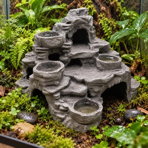 Peut inclure: Une formation rocheuse grise à plusieurs niveaux pour terrariums, dotée de plusieurs bassins circulaires intégrés pour l'eau ou la nourriture, de multiples entrées de grottes en arc et d'une surface texturée imitant la pierre naturelle.