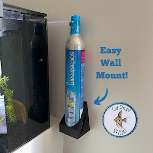 以下が含まれることがあります： ソーダストリームのCO2シリンダー用の黒い壁掛けマウント。マウントは水槽の隣にある壁に取り付けられています。壁には「Easy Wall Mount!」という文字が書かれています。青い銀色のソーダストリームCO2シリンダーがマウントに収められています。シリンダーには「sodastream」という文字が書かれています。画像の右下には、「Lid Propz SHOP」という文字が書かれた青い円があります。