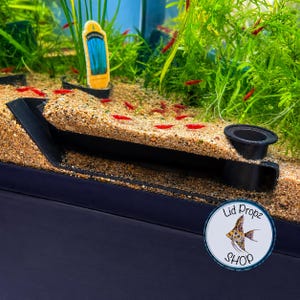 May include: A black plastic aquarium lid prop featuring a rectangular frame with a circular opening on one side, partially buried in light brown sand within a fish tank. The prop is positioned near green aquatic plants and small red shrimp.