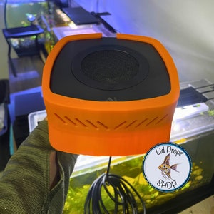 Op de afbeelding: Oranje en zwarte aquariumluchtpomp met een rond luidsprekerrooster en een snoer. De pomp wordt in een hand met een groene mouw vastgehouden. De tekst "Lid Propz Shop" is zichtbaar in een cirkel met een visafbeelding.