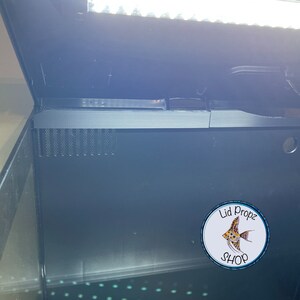 May include: A black aquarium tank with a black lid and a white light fixture. The tank is empty and has a black grate on the bottom. A sticker with the text "Lid Propz Shop" and a fish design is on the side of the tank.