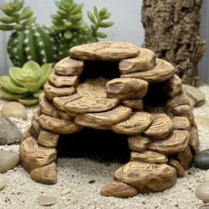 May include: A dome-shaped reptile hideout constructed from stacked, tan-colored stone-like segments. The structure features a large arched entrance at the front and a smaller side opening, creating a hollow interior space for small animals.