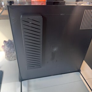 May include: A black aquarium with a glass front. The top of the aquarium has a black mesh cover. A black plastic piece with angled slots is attached to the side. The word "MAX" is printed on the side.