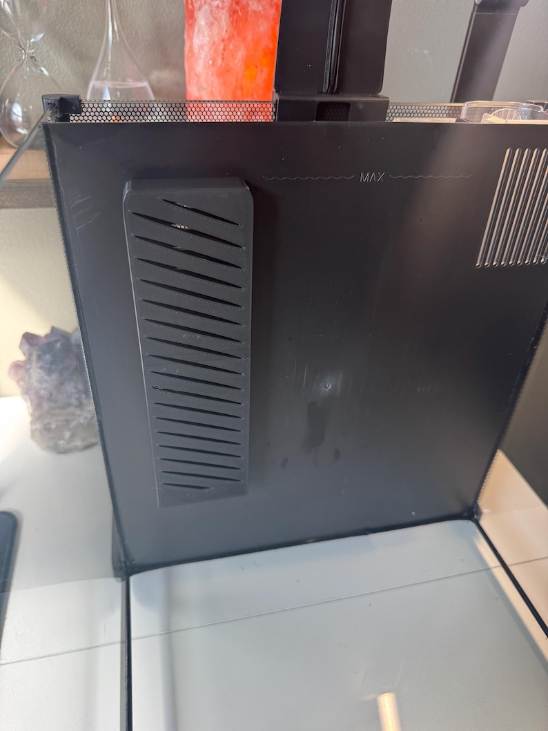 May include: A black aquarium with a glass front. The top of the aquarium has a black mesh cover. A black plastic piece with angled slots is attached to the side. The word "MAX" is printed on the side.