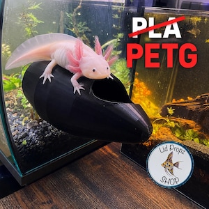 Peut inclure: Un axolotl rose clair repose sur une cachette d'aquarium cylindrique noire et creuse. Le texte PLA est barré d'une ligne rouge au-dessus du texte rouge PETG. Un logo circulaire avec une illustration de poisson et le texte Lid Propz SHOP.