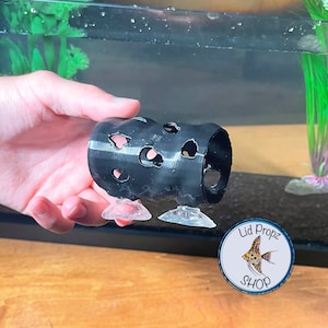 May include: Black plastic fish tank cave with multiple holes and two suction cups. The cave is being held in a hand over a fish tank. The text "Lid Propz Shop" is visible in a circle with a fish graphic.