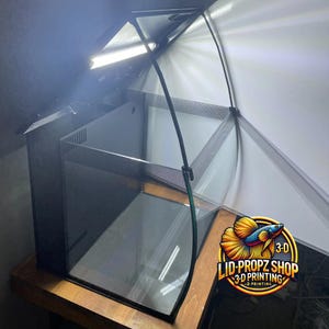 May include: A black curved glass aquarium with a black lid and a black mesh divider. The aquarium is on a wooden surface. The lid is open and has a white light inside. The aquarium has a black and white logo on the front that says "LID-PROPZ SHOP 3-D PRINTING".