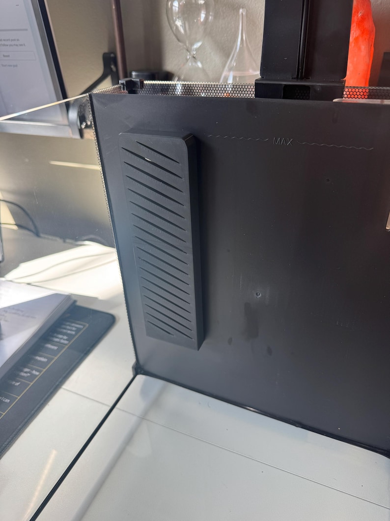 May include: A black computer monitor with a black, rectangular, ridged component attached to the side. The monitor has a maximum level indicator. The monitor is on a white surface.