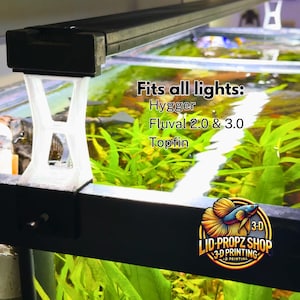 May include: A white 3D printed aquarium light fixture bracket. The bracket is designed to fit Hygger, Fluval 2.0 & 3.0, and Topfin aquarium lights. The text "Fits all lights: Hygger Fluval 2.0 & 3.0 Topfin" is visible in the image. The bracket is attached to a black aquarium.