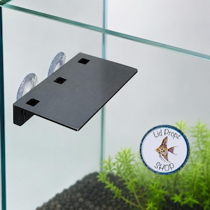 Puede incluir: Un estante rectangular negro para acuario con tres aberturas cuadradas, fijado a la pared de cristal de un tanque mediante dos ventosas transparentes. Se ve una pegatina circular con el texto Lid Propz SHOP y un pez ángel ilustrado.
