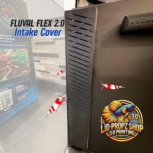 以下が含まれることがあります： 黒色のFluval Flex 2.0吸入口カバー。斜めのスロットと青色の照明があります。「FLUVAL FLEX 2.0 Intake Cover」の文字が見えます。画像には、3D Lid-Propz Shopのロゴと、赤と白のエビのイラストも含まれています。
