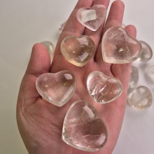 Könnte beinhalten: Eine Sammlung herzförmiger Steine aus klarem Quarz. Die Steine variieren in Größe und Klarheit, wobei einige durchscheinender erscheinen als andere. Die Herzen werden in einer Hand gehalten, wodurch ihre glatten, polierten Oberflächen hervorgehoben werden. Der Gesamteindruck ist von zarter Schönheit.