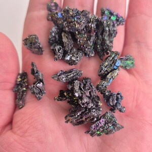 May include: A collection of iridescent silicon carbide crystals, displaying a range of colors including purple, green, and blue. The crystals have a rough, textured surface and are held in a hand.