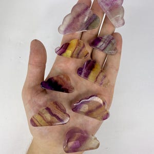 May include: A collection of cloud-shaped fluorite crystals in shades of purple, yellow, and clear. The polished stones are held in a hand against a white background. The crystals have a translucent quality, showcasing the natural color variations.