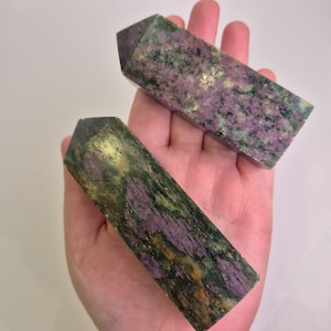 Puede incluir: Dos obeliscos rectangulares de piedras preciosas pulidas. Las piedras presentan una mezcla de colores, incluyendo púrpura intenso, verde y dorado. Los obeliscos se sostienen en una mano, mostrando su tamaño y forma.