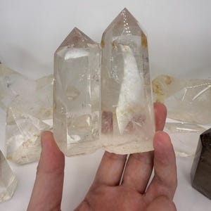 May include: Two clear quartz crystal points with natural inclusions. The crystals are held in a hand, with other crystals visible in the background. The crystals have a pointed top and a rough base. The crystals are a light, translucent color.