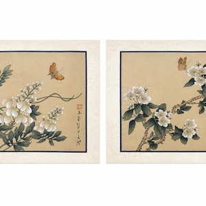 May include: Two rectangular art prints featuring botanical illustrations on a tan background. The left print shows white flowers, green leaves, and an orange butterfly. The right print displays white blossoms, green foliage, and a butterfly, with red stamps and black calligraphy.