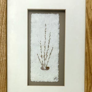 Può includere: Un'opera d'arte incorniciata con un pezzo verticale di carta bianca fatta a mano, testurizzata e con bordi irregolari. La carta mostra un'illustrazione a inchiostro marrone di rami sottili con gemme che emergono da una piccola base. Firma: "Amhu 99".
