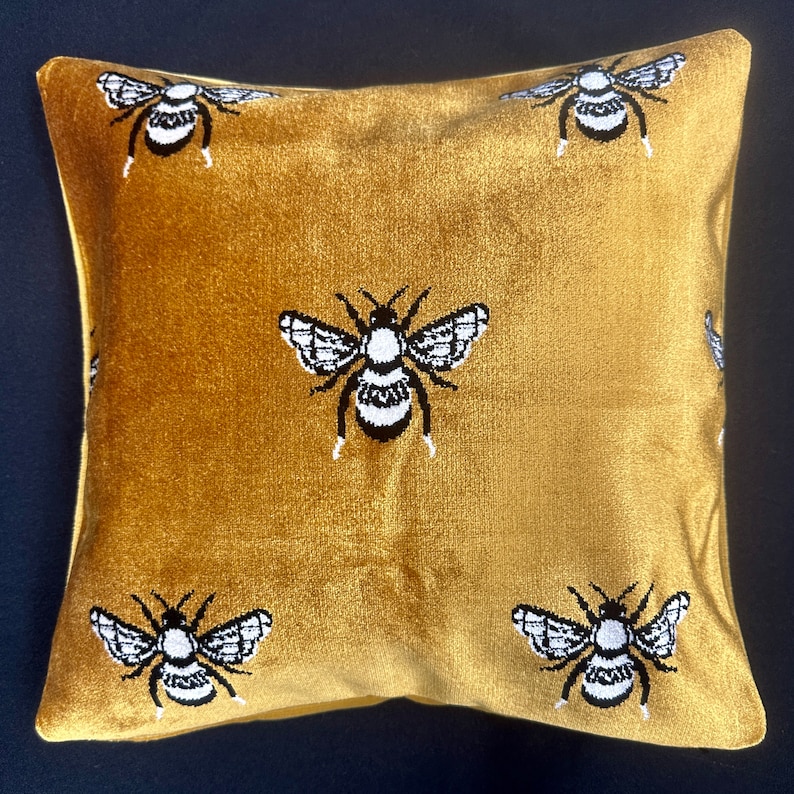 May include: A gold velvet pillow with a repeating pattern of black and white embroidered bees.