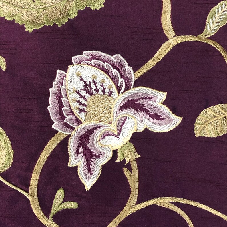 May include: Purple fabric with an embroidered floral design in shades of white, green, and gold. The fabric features a large flower with a white centre and purple petals.