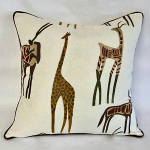 Könnte beinhalten: Ein weißes Kissen mit braunem Rand zeigt ein gesticktes Design von Giraffen, Zebras und Antilopen in Braun, Grün und Gold.