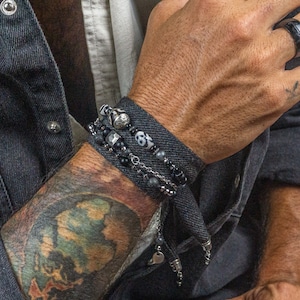 Könnte beinhalten: Nahaufnahme eines Arms mit mehreren schwarzen Armbändern mit Totenkopf- und Perlendetails. Ein schwarzer Ring ist an der Hand zu sehen. Der Arm ist tätowiert. Der Hintergrund ist eine Backsteinmauer. Die Armbänder sind aus Metall und Perlen.
