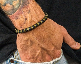 May include: A black and gold beaded bracelet on a wrist with a colorful tattoo of a world map. The person is wearing a black long-sleeve shirt and light blue distressed jeans. The hand is in the pocket of the jeans.