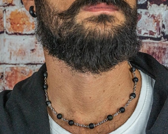 May include: A close-up of a man wearing a black hooded sweatshirt and a white t-shirt. He is wearing a black beaded necklace with silver accents.