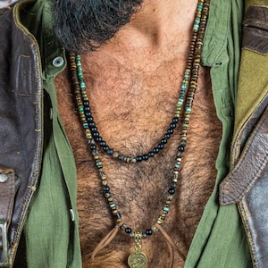 May include: A layered necklace with black, brown, and turquoise beads, and a pendant. The necklace is worn over a green shirt and a distressed brown leather jacket. The jacket has a vintage look with visible wear and tear.