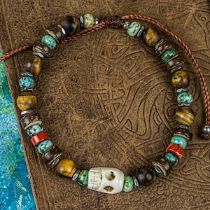 May include: A beaded bracelet with a skull charm. The bracelet features a variety of beads in colors like brown, turquoise, red, and yellow. The bracelet is on a brown leather surface.