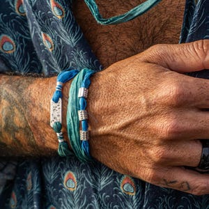 May include: Close-up of a person's arm and hand with jewellery. Two blue bracelets with silver accents and a black ring are visible. The person is wearing a blue shirt with a peacock feather pattern.
