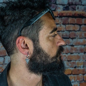 May include: A man with a beard and curly hair is wearing a black jacket and sunglasses. He has a silver hoop earring with a triangle pendant in his left ear.