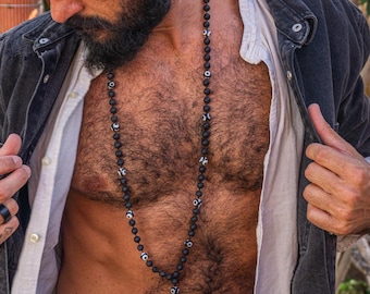 May include: A man wearing a black denim jacket over a white shirt, with a black beaded necklace with silver accents. The necklace is visible through the open jacket.