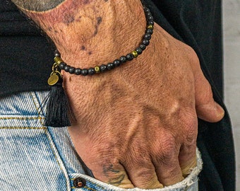 May include: A black beaded bracelet with gold-colored accents and a black tassel charm. The bracelet is worn on a wrist, paired with a black long-sleeve shirt and light blue jeans. The image is set against a white brick wall.